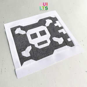 Könnte beinhalten: Ein handgefertigter Quiltblock mit einem Totenkopf-Design. Der Totenkopf ist weiß auf einem dunkelgrauen Hintergrund, mit einem weißen Rand. Der Text "QUILTS and NICE STUFF" befindet sich in der oberen rechten Ecke.
