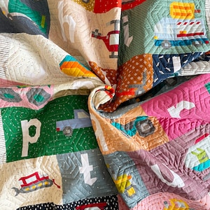 May include: A colorful patchwork quilt featuring various construction vehicles and the letters of the alphabet. The quilt is made of squares in shades of red, green, blue, and yellow. The quilt is approximately 60 inches by 70 inches.