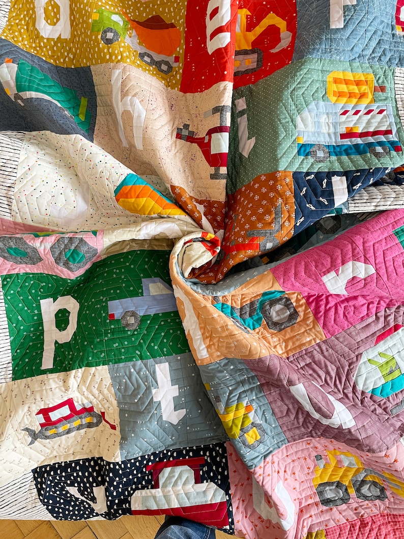 May include: A colorful patchwork quilt featuring various construction vehicles and the letters of the alphabet. The quilt is made of squares in shades of red, green, blue, and yellow. The quilt is approximately 60 inches by 70 inches.