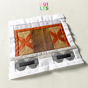 Könnte beinhalten: Ein handgefertigter Quiltblock mit einem zentralen Design aus orangefarbenen Sternen und braunem Stoff, umrahmt von einem weißen Rand. Der untere Abschnitt hat zwei geometrische Formen in Schwarz und Grau. Der Text "QUILTS AND NICE STUFF" befindet sich in der oberen rechten Ecke.