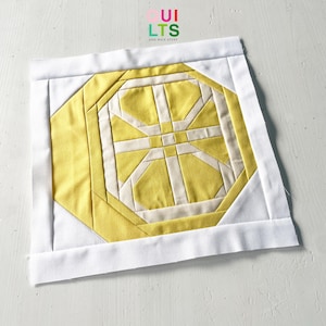 Könnte beinhalten: Ein quadratischer, handgefertigter Quiltblock mit einem geometrischen Design in Gelb- und Cremetönen, umrahmt von einem weißen Rand. Das zentrale Muster ist ein stilisiertes, symmetrisches Design. Der Text "QUILTS" und "AND NICE STUFF" ist sichtbar.