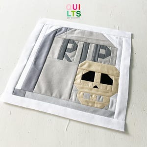 Könnte beinhalten: Ein quadratischer Stoff-Quiltblock mit einem Totenkopf-Design. Der Schädel ist beige mit schwarzen Augenhöhlen und einer kleinen dreieckigen Nase. Der Hintergrund ist grau, mit einem weißen Rand. Der Text "QUILTS AND NICE STUFF" befindet sich in der oberen rechten Ecke.