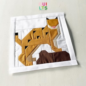 Könnte beinhalten: Ein geometrischer Patchwork-Quiltblock mit einer stilisierten braunen und braunen Katze mit rosa Nase. Der Block ist mit einem weißen Rand umrahmt.