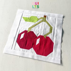 Może przedstawiać: Biały kwadrat materiału z geometrycznym wzorem dwóch czerwonych wiśni, zielonych liści i brązowej łodygi. Wzór wykonany jest z zszytych ze sobą kawałków materiału. Tekst "QUILTS AND NICE STUFF" znajduje się w prawym górnym rogu.