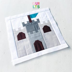 Puede incluir: Un bloque de edredón de tela cuadrado con un diseño de castillo hecho de piezas en tonos gris y marrón oscuro, rematado con una bandera verde azulado. El castillo está enmarcado por un borde blanco y el texto QUILTS AND NICE STUFF arriba.