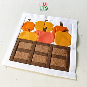 May include: A square fabric quilt block featuring a pieced design of orange and yellow pumpkins sitting in a brown wooden crate. The block is framed by a white border and includes the text QUILTS AND NICE STUFF at the top centre.