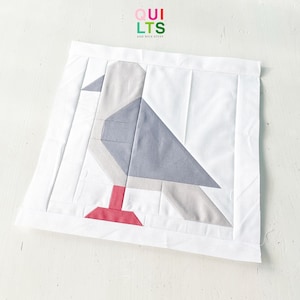 May include: A square, white quilt block featuring a geometric bird design. The bird is composed of gray, light gray, and red fabric pieces. The block is bordered by a white fabric frame. The text "QUILTS" and "AND NICE STUFF" is visible in the upper right corner.