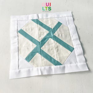 May include: A white fabric square with a teal geometric pattern in the center. The fabric square is surrounded by a white border. The text "QUI LTS AND NICE STUFF" is printed in the top center of the image.