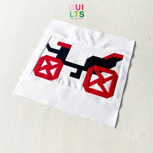 Könnte beinhalten: Ein quadratischer Stoff-Quiltblock mit einem zusammengesetzten Design eines Fahrrads in Schwarz, Rot und Weiß. Das Fahrrad hat zwei rote Speichenräder, einen schwarzen Rahmen und einen roten Sattel. Der Hintergrund ist aus weißem Stoff. Der Text QUILTS AND NICE STUFF ist sichtbar.