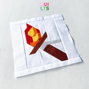 May include: A square fabric quilt block featuring a pieced design of a flaming torch. The torch has a brown handle and a flame composed of yellow, orange, and red fabric segments. The background is white fabric. The logo QUILTS AND NICE STUFF is visible.