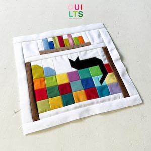 May include: A square fabric quilt block featuring a pieced design of a black cat silhouette sitting on a colorful grid of small squares. Above the cat is a brown shelf holding vertical fabric strips in blue, green, red, and orange colors.