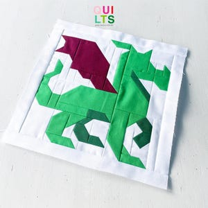 May include: A square fabric quilt block featuring a pieced dragon design in shades of bright green and dark green with a deep maroon wing. The dragon is centered on a white background with a white border. The text QUILTS AND NICE STUFF is visible.