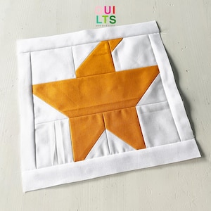 Könnte beinhalten: Ein weißer Stoffquadrat mit einem großen, gelben Stern in der Mitte. Der Stern besteht aus mehreren Stoffteilen, die zusammengenäht sind. Der Stoffquadrat hat einen weißen Rand.