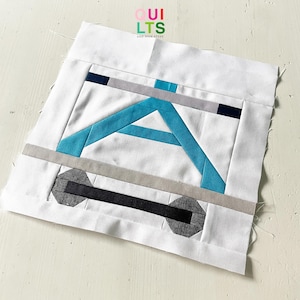 May include: A white fabric quilt block with a geometric design. The design features a large teal letter "A", gray and black accents, and the text "QUILTS AND NICE STUFF" in the upper right corner.