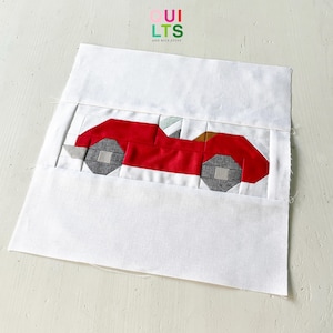 May include: A handmade quilt block featuring a red car design. The car has gray wheels and is set against a white background. The text "QUILTS AND NICE STUFF" is visible in the upper right corner.