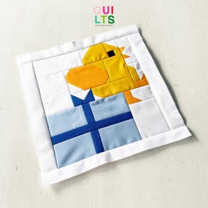 May include: A patchwork quilt square with a yellow duck and a blue gift. The quilt has a white border and the text "QUILTS AND NICE STUFF". The duck has an orange beak.