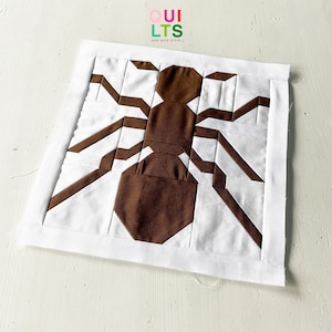 Könnte beinhalten: Ein brauner und weißer Patchwork-Quiltblock mit einem geometrischen Spinnen-Design. Der Quiltblock besteht aus braunen und weißen Stoffquadraten, die zusammengenäht wurden, um die Spinnenform zu erzeugen.