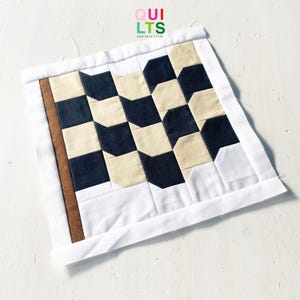 May include: A handmade quilt square with a geometric pattern of black, beige, and white shapes. The quilt has a white border and a brown strip on the left side. The text "QUILTS AND NICE STUFF" is at the top.