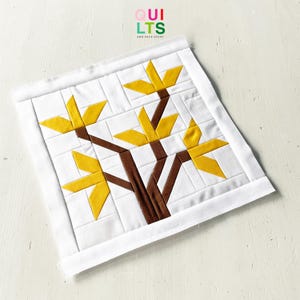 May include: A white quilted square featuring a tree design. The tree has brown branches and yellow leaves. The text "QUILTS AND NICE STUFF" is in the upper right corner. The quilt is bordered with white fabric.