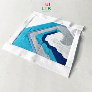 May include: A blue and gray geometric quilt block with a white border. The block is made up of various shapes and sizes of fabric, creating a modern and abstract design.