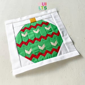 May include: A square, handmade Christmas ornament quilt block. The design features a green ornament with red and white accents, framed by a white border. The ornament has a gold top. The text "QUILTS AND NICE STUFF" is in the upper right corner.