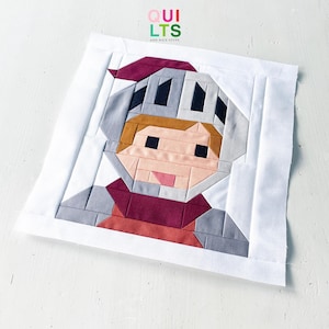 Op de afbeelding: Een vierkant stoffen quiltblok met een samengesteld ontwerp van een ridder die een grijze helm met een kastanjebruine pluim draagt. Het figuur heeft een getinte huid, bruin haar en een kastanjebruine kraag over een grijs harnas, omlijst door een witte rand met de tekst QUILTS AND NICE STUFF.