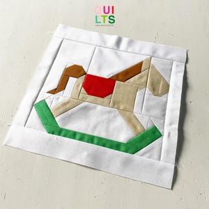 May include: A fabric quilt block featuring a patchwork rocking horse design. The horse is constructed from tan, brown, and cream-colored fabric with a red saddle and green rockers, set against a white background. The block is framed with a white border.