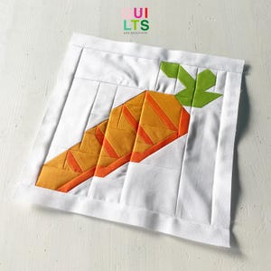 Könnte beinhalten: Ein weißes Stoffquadrat mit einem Patchwork-Karotten-Design. Die Karotte ist orange mit orangefarbenen Streifen und grünen Blättern. Der Text "QUILTS AND NICE STUFF" befindet sich in der oberen rechten Ecke.