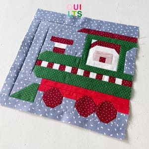 May include: A green and red patchwork train block with a white roof and a pink window. The block is on a blue background with white polka dots.