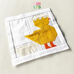May include: A patchwork quilt block with a geometric design of a yellow chick emerging from an egg. The chick has orange feet and a white and yellow crown. The block is framed with a white border. The text "QUILTS AND NICE STUFF" is in the upper right corner.