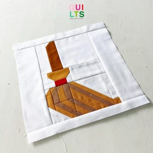 Könnte beinhalten: Ein handgefertigter Quiltblock mit einem Besen-Design. Der Besen besteht aus verschiedenen Brauntönen mit einem roten Akzent. Der Block ist vor einem weißen Hintergrund platziert, mit dem Text "QUILTS AND NICE STUFF" oben rechts.