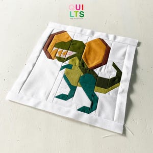 May include: A white fabric square with a green, brown, and yellow dinosaur applique. The dinosaur is made of fabric pieces sewn together. The fabric square has a white border and the text "OUI LTS AND NICE STUFF" is printed in the top centre.
