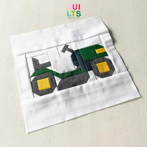 May include: A fabric quilt block featuring a tractor design. The tractor is composed of green, gray, white, and yellow fabric pieces. The block is framed by a white border, and the text "QUILTS AND NICE STUFF" is visible in the upper left corner.