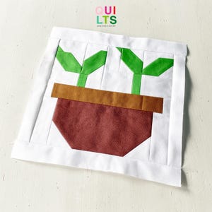 May include: A square quilt block with a geometric design of a potted plant. The pot is a deep red-brown, with green leaves and a brown rim. The background is white, with the text "QUILTS AND NICE STUFF" in the upper right corner.