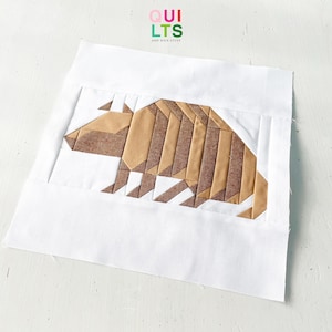 May include: A white fabric square with a geometric armadillo design in shades of brown and tan. The armadillo is centered on the square, with the text "QUILTS AND NICE STUFF" in the upper right corner.