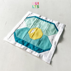 May include: A teal, blue, and yellow geometric quilt block with a white border. The quilt block is made of fabric squares sewn together to create a geometric pattern. The quilt block is on a white surface.