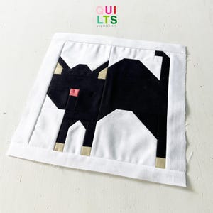 Könnte beinhalten: Ein Stoffquadrat mit einem geometrischen Schwarz-Katzen-Design. Die Katze befindet sich vor einem weißen Hintergrund, mit einer rosa Nase und beigen Akzenten an Pfoten und Ohren. Die Worte "QUILTS" und "NICE STUFF" sind oben rechts aufgedruckt.