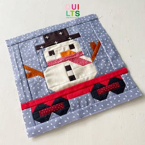 May include: A patchwork quilt block featuring a snowman with a black hat, red scarf, and orange carrot nose. The block is set against a blue background with white polka dots. The block is surrounded by a red, black, and white border.