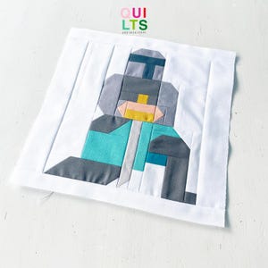 Può includere: Un blocco di trapunta in tessuto quadrato con un design a pezzi di una figura corazzata stilizzata in tonalità grigio scuro, grigio chiaro, verde acqua e giallo, incorniciato da un bordo bianco. Il testo QUILTS AND NICE STUFF è stampato in alto.