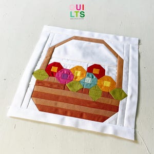 May include: A square fabric quilt block featuring a pieced basket design with a brown handle and base. The basket is filled with colorful geometric flowers in red, pink, yellow, blue, and orange, accented by green leaves on a white background.