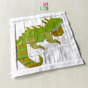 May include: A green and yellow geometric quilt block featuring a stylised iguana design. The quilt block is on a white background and has a white border.