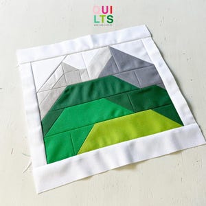 May include: A square fabric quilt block featuring a mountain landscape design. The scene includes grey and white peaks above layered green hills in varying shades of lime, forest, and emerald green, all framed by a solid white fabric border.