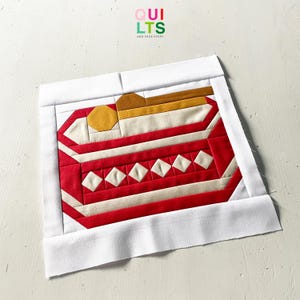 May include: A quilted textile with a geometric design in red, white, and gold. The central pattern features a series of red and white stripes, with a row of white diamond shapes. The piece is framed by a white border. The text "QUILTS AND NICE STUFF" is visible.