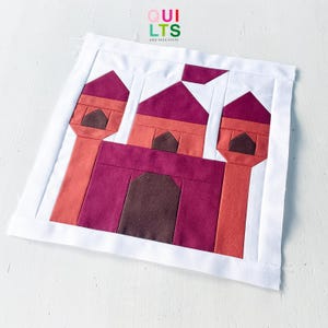 May include: A square fabric quilt block featuring a pieced castle design in shades of magenta, coral, and dark brown, framed by a white border. The castle includes three towers with pointed roofs and a central doorway, with the text QUILTS AND NICE STUFF printed above.