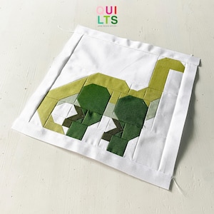 Könnte beinhalten: Ein weißes Stoffpanel mit einer grünen und gelben Dinosaurier-Applikation. Der Dinosaurier besteht aus zusammengenähten Stoffquadraten. Das Panel trägt oben den Text "OUI LTSS AND NICE STUFF".