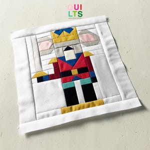 Puede incluir: Un bloque de tela colorido con un diseño de cascanueces. El cascanueces tiene una chaqueta roja, pantalones negros y una corona dorada. El bloque está enmarcado en tela blanca, con el texto "QUILTS AND NICE STUFF" en la esquina superior derecha.