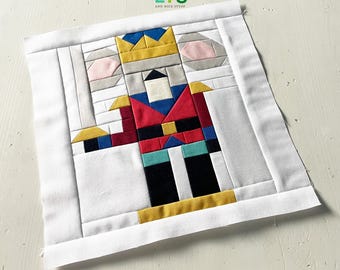 May include: A colorful fabric block featuring a nutcracker design. The nutcracker is depicted with a red jacket, black pants, and a gold crown. The block is framed in white fabric, with the text "QUILTS AND NICE STUFF" in the upper right corner.