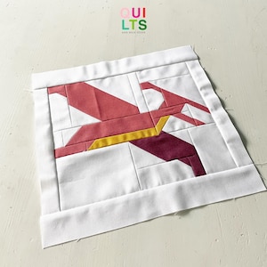 May include: A white quilt block with a geometric design featuring red, burgundy, and yellow fabric pieces. The block is surrounded by a white border.