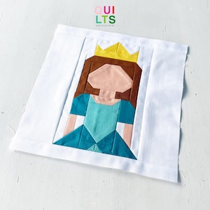 Op de afbeelding: Een vierkant stoffen quiltblok met een samengesteld ontwerp van een figuur met bruin haar, een gele kroon en een blauwgroene jurk op een witte achtergrond. De tekst QUILTS AND NICE STUFF staat in kleine letters boven het centrale figuur.