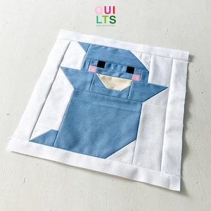 Könnte beinhalten: Ein quadratischer, handgefertigter Quiltblock mit einem blauen Cartoon-Charakter mit weißem Bart und weißem Rand. Der Charakter hat schwarze Augen, rosa Wangen und einen cremefarbenen Mund. Der Text "QUILTS AND NICE STUFF" befindet sich oben rechts.